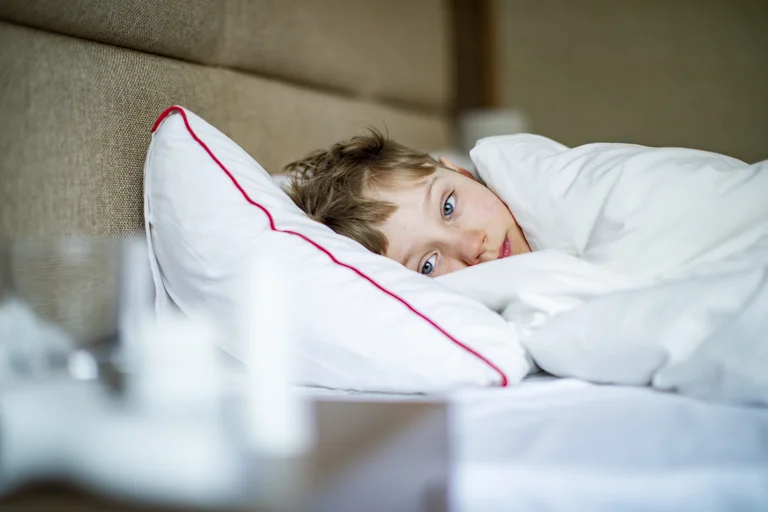 Krankes Kind liegt im Bett, zugedeckt mit einer Decke und zeigt einen besorgten Blick.