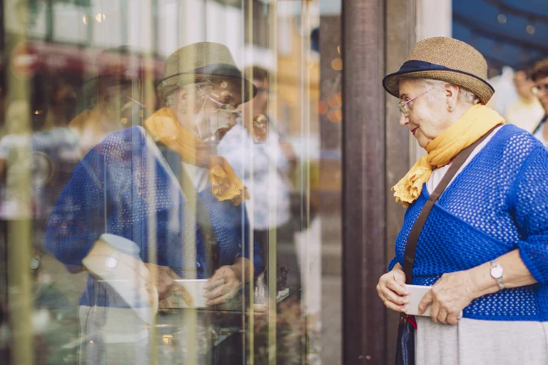 Ältere Frau steht vor einem Schaufenster und betrachtet Schuhe, während sich ihre Spiegelung im Glas zeigt. 
