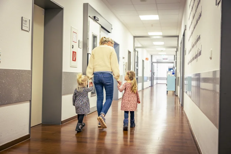 Zwei Kinder gehen Hand in Hand mit ihrer Mutter über den Flur eines Krankenhauses.