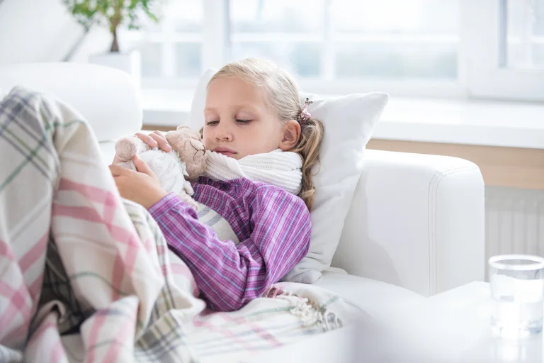 Krankes Mädchen ruht sich mit einem Stofftier auf der Couch aus, eingehüllt in eine Decke.