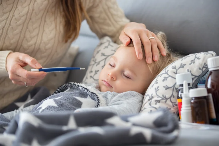 Kind liegt mit Fieber auf der Couch. Mutter fühlt mit einer Hand die Temperatur an der Stirn und hält in der anderen ein Thermometer.