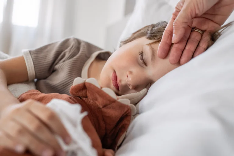 Kind liegt schlafend im Bett, Erwachsener prüft mit der Hand die Körpertemperatur an der Stirn.