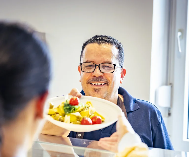 Ein Mitarbeiter der Cafeteria reicht einer Besucherin ein Nudelgericht an