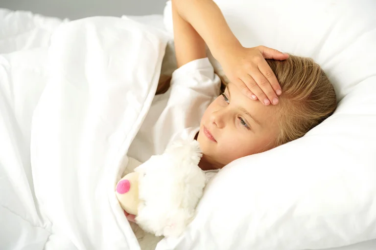 Krankes Kind liegt mit Stofftier im Bett und hält sich die Stirn.