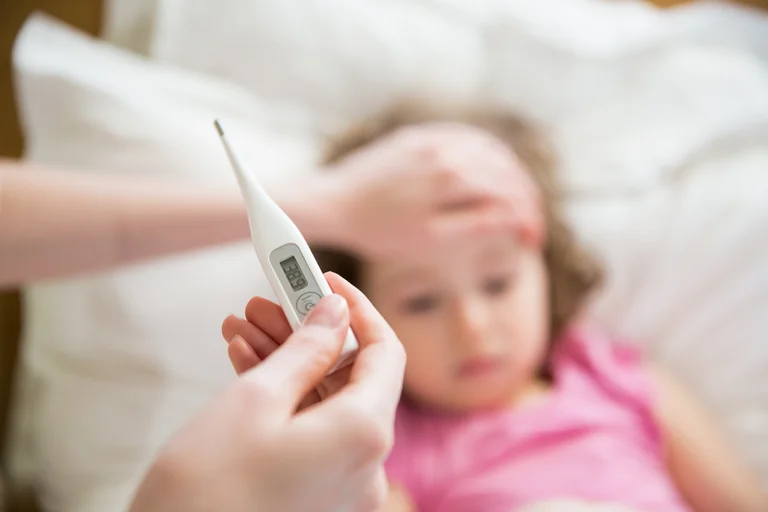 Fieberthermometer mit krankem Kind im Hintergrund, dessen Temperatur and er Stirn geprüft wird.