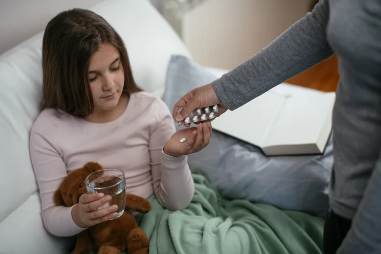 Krankes Mädchen im Bett nimmt Tabletten aus der Hand einer Betreuungsperson, während es ein Stofftier hält und ein Glas Wasser in der Hand hat.